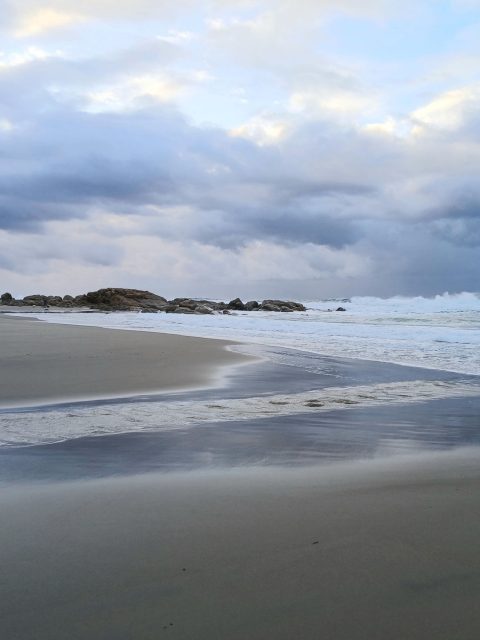 Wet sand, foam and dark rocks under an overcast sky